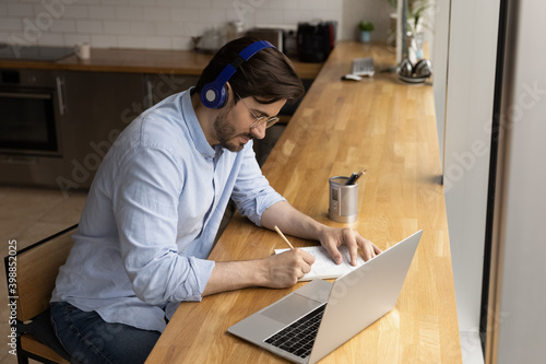 Top view of millennial man in earphones study or work online on computer making notes. Young Caucasian male in headphones take distant course on laptop writing and summarizing. Education concept.