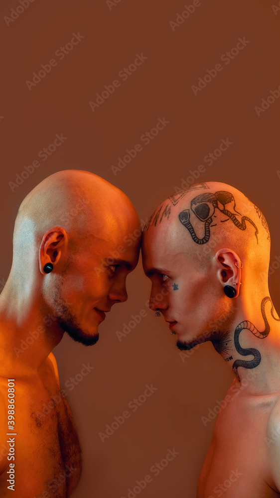 Portrait of young twin brothers with tattoos and piercings standing head to head, looking at each other isolated over orange background Portrait of young twin brothers with tattoos and piercings standing head to head, looking at each other isolated over orange background