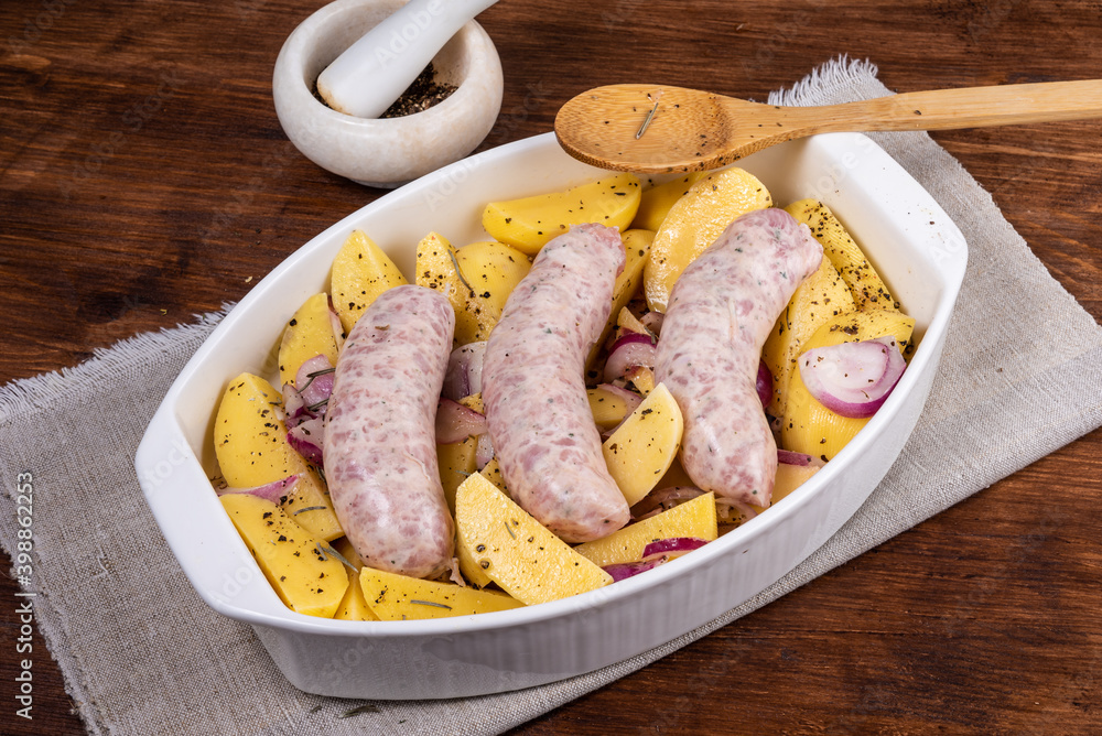 Sliced raw potatoes with onions with black pepper and sausages in a baking dish on a wooden table, top view Sliced raw potatoes with onions with black pepper and sausages in a baking dish on a wooden table, top view