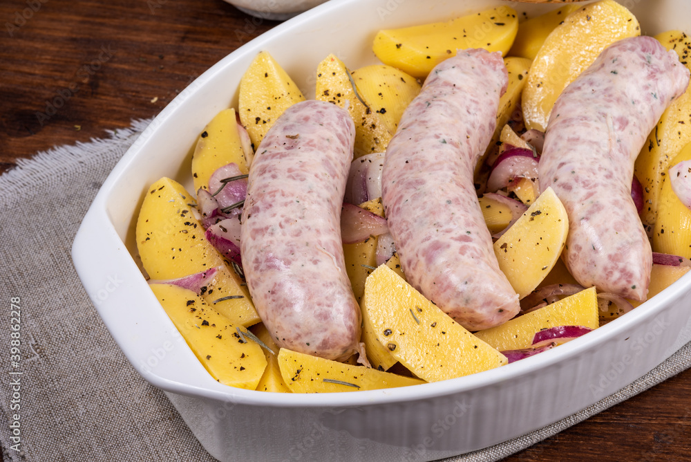 Sliced raw potatoes with onions with black pepper and sausages in a baking dish on a wooden table, top view, close-up Sliced raw potatoes with onions with black pepper and sausages in a baking dish on a wooden table, top view, close-up