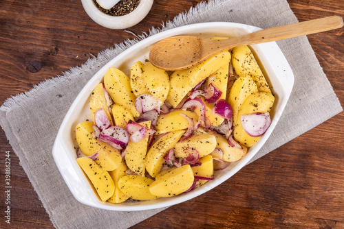 Sliced raw potatoes with onions with black pepper and sausages in a baking dish on a wooden table, top view