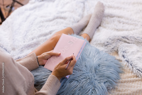 Top view of woman in wool sock and sweater with pink notebook sign 2021, large tattoo on hip. Female sit on bed at home in bedroom.   