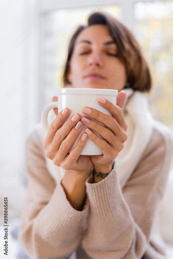Warm portrait of woman sitting on windowsill with cup of hot tea coffee wearing sweater and white scarf, warm herself in apartment, cold weater outside. Warm portrait of woman sitting on windowsill with cup of hot tea coffee wearing sweater and white scarf, warm herself in apartment, cold weater outside.