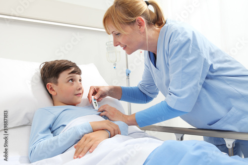 Happy child lying in bed in hospital room and nurse taking his temperature 