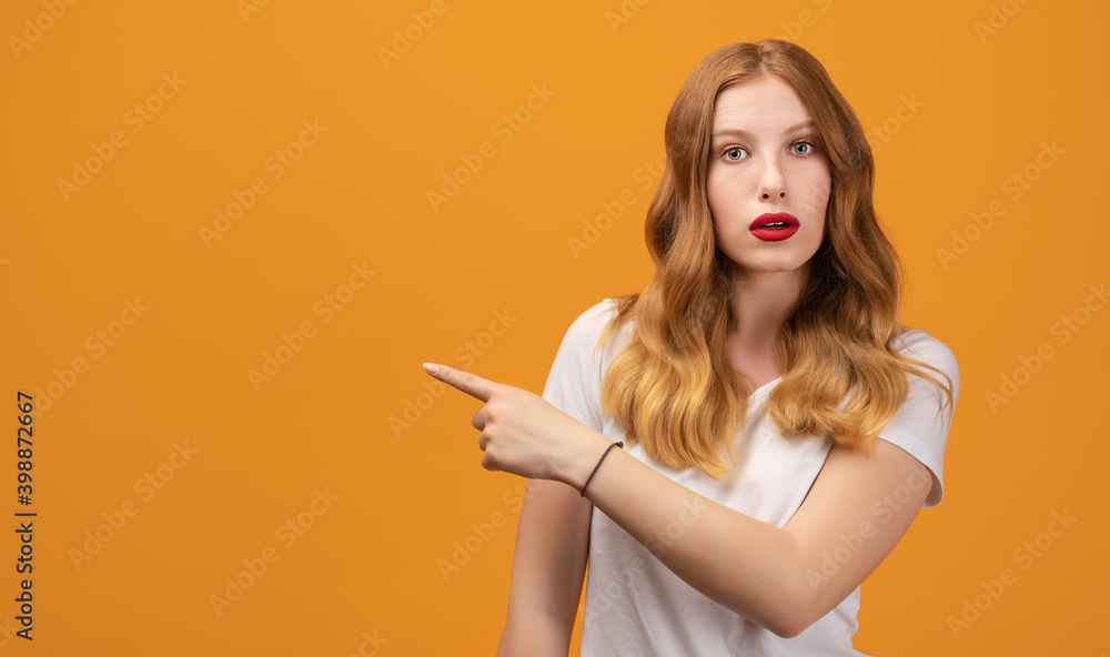 Pretty girl with wavy redhead surprised and pointing finger to the side, shows copy space for your information