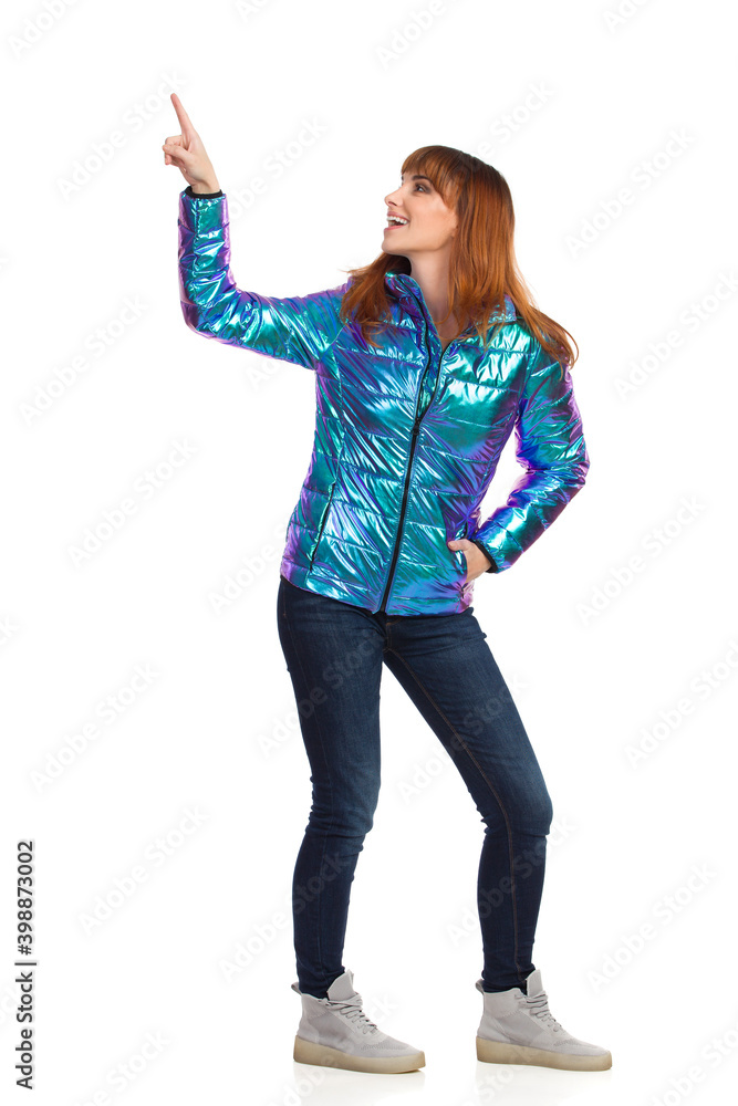 Smiling Young Woman In Vibrant Down Jacket Is Looking Back And Pointing Up Smiling Young Woman In Vibrant Down Jacket Is Looking Back And Pointing Up
