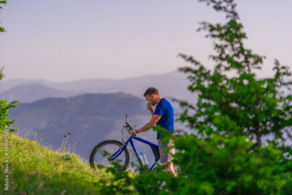 Fototapeta premium Tired cyclist is wiping his sweat off his face while pushing his bicycle uphill on a dirt road in a mountain.