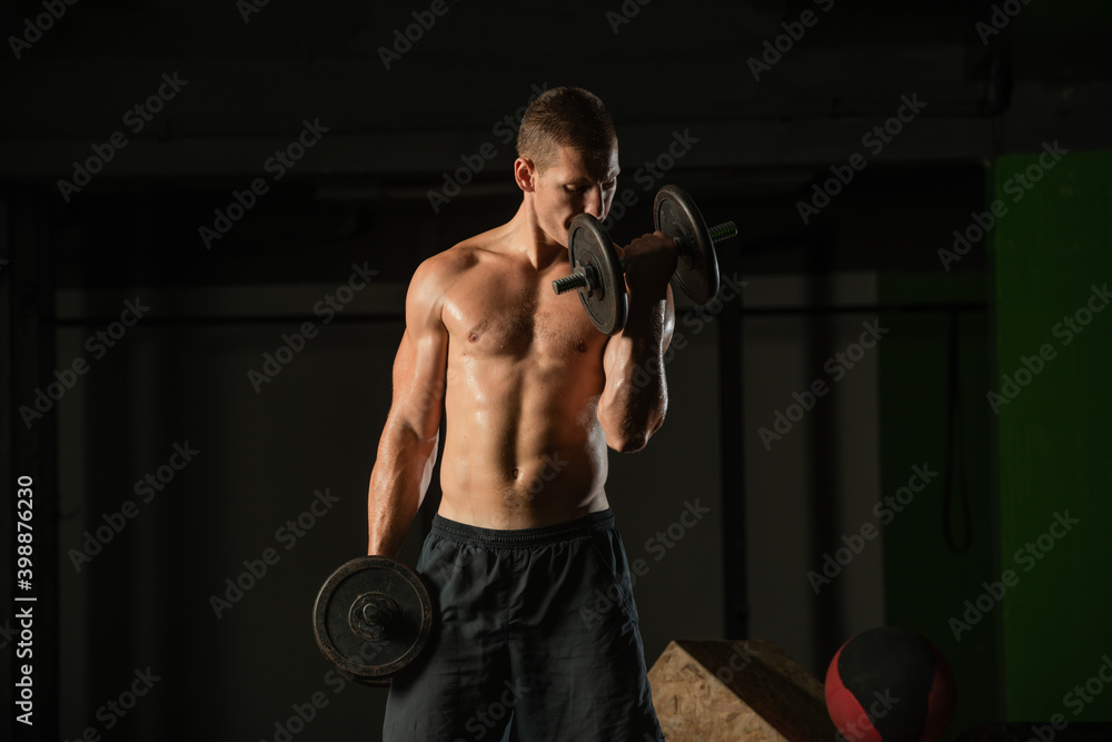 Close up dark portrait of a shirtless young man exercising dumbbell alternate biceps curl