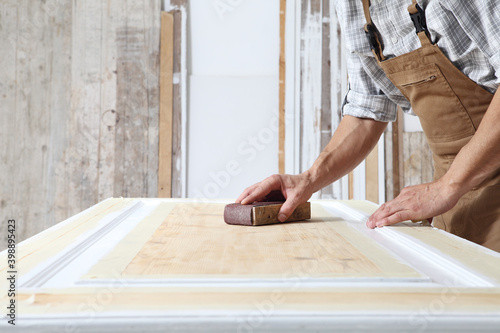 Male carpenter working the wood in carpentry workshop, sanding a wooden door with sandpaper, wearing overall