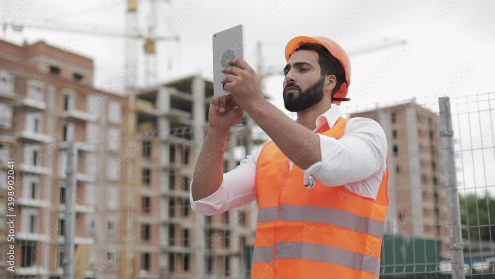 Portrait of a successful young businessman, wearing a white helmet, in a suit working with tablet looking at the camera. The builder stands against the backdrop of a modern building. Portrait of a successful young businessman, wearing a white helmet, in a suit working with tablet looking at the camera. The builder stands against the backdrop of a modern building.