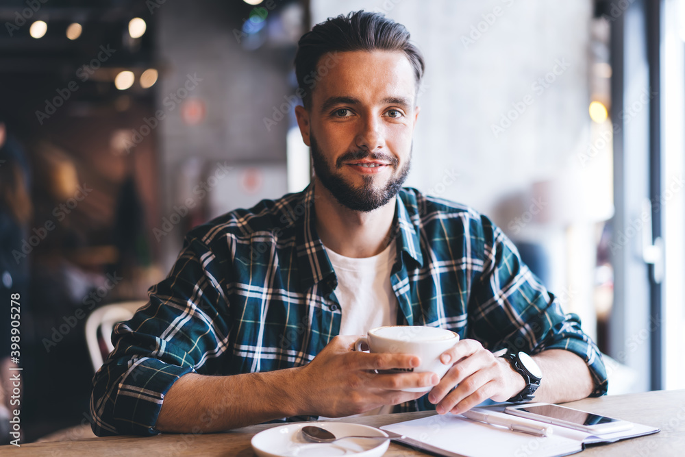 Portrait of happy male writer with textbook smiling at camera spending daytime for creating idea in cafe interior, successful hipster guy with coffee cup sitting at cafeteria table and posing Portrait of happy male writer with textbook smiling at camera spending daytime for creating idea in cafe interior, successful hipster guy with coffee cup sitting at cafeteria table and posing