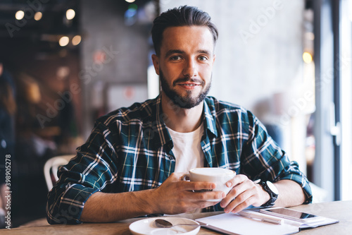 Portrait of happy male writer with textbook smiling at camera spending daytime for creating idea in cafe interior, successful hipster guy with coffee cup sitting at cafeteria table and posing