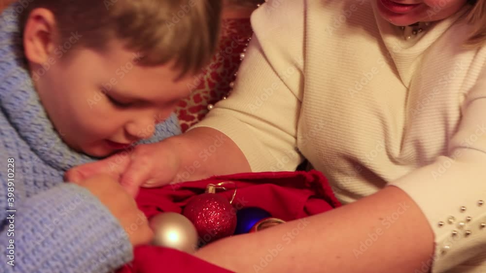 Close-up boy bent over and examines Christmas balls in a bag. Beautiful toys on a coniferous garland. Against the background of a log wall of a wooden house. We celebrate the new year. Warm light. 4K. Close-up boy bent over and examines Christmas balls in a bag. Beautiful toys on a coniferous garland. Against the background of a log wall of a wooden house. We celebrate the new year. Warm light. 4K.