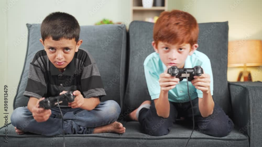 Two diverse kids seriously busy playing video game using joystick at home while sitting on sofa - concept of kids game concentration and leisure activity