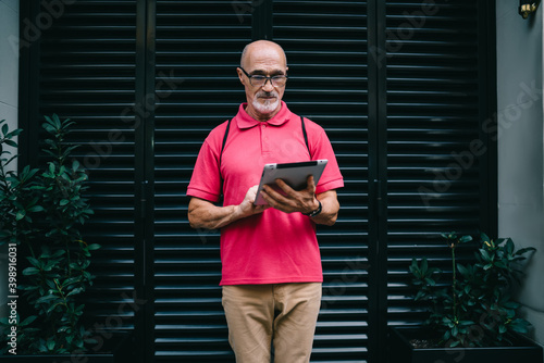 Elderly blogger connecting to 4g wireless for chatting and messaging in online mode, senior in classic eyeglasses reading received email on modern touch pad standing at urbanity and networking