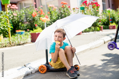 Cheerful boy in a blue t-shirt sits on a skateboard under a white umbrella