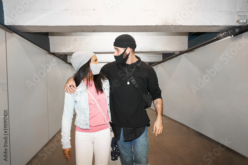 Young interracial couple of lovers in an underground subway corridor wearing face masks walking in each other's arms down an old public transportation corridor and looking at each other.