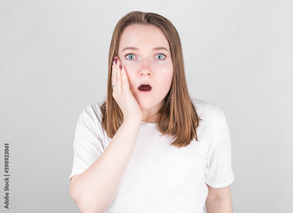 Young woman in a white T-shirt stands against a gray wall and holding on to face in great surprise. Young woman in a white T-shirt stands against a gray wall and holding on to face in great surprise.