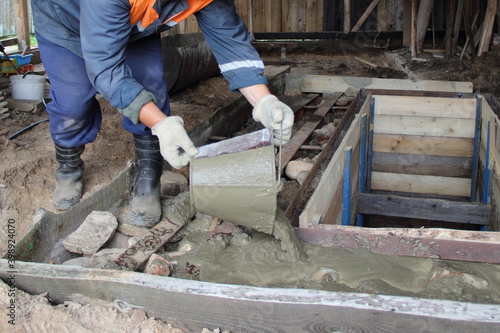 Working man in overalls pours concrete from a bucket on the floor around the garage observation pit, independent village indoor industrial construction building © Ilya