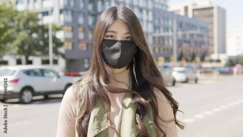 A young Asian woman in a face mask looks at the camera in a street in an urban area - cars drive on the busy road in the blurry background A young Asian woman in a face mask looks at the camera in a street in an urban area - cars drive on the busy road in the blurry background