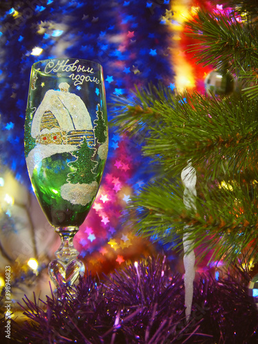 Green glass with new-year pictire near a fragment of new-year tree; colored bokeh as the background photo
