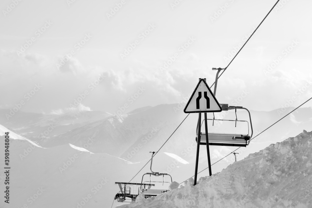 Warning traffic sign road narrows, chairlift and high snowy mountains in fog Warning traffic sign road narrows, chairlift and high snowy mountains in fog