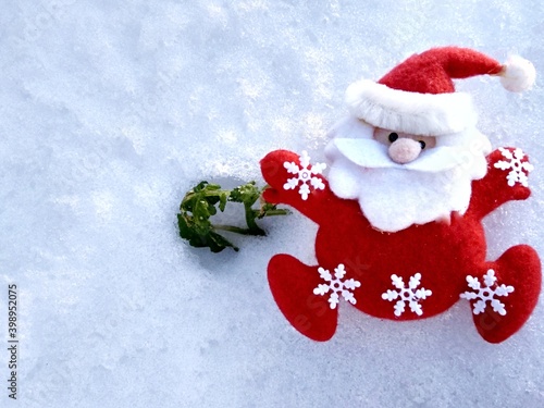 santa toy in red clothes with white snowflakes on a snowy background with green leaves