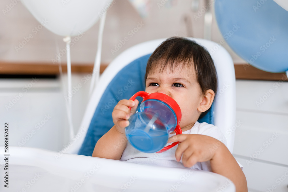 Little baby boy sitting in blue high chair at home on white kitchen and drinking water from sippy cup on background with balloons. Little baby boy sitting in blue high chair at home on white kitchen and drinking water from sippy cup on background with balloons.