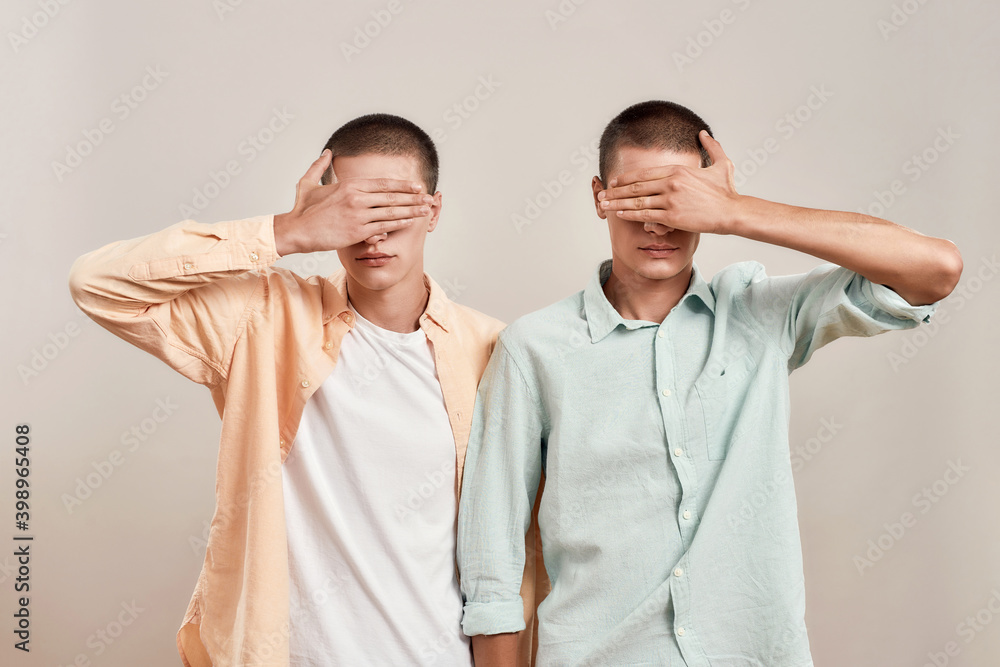 Portrait of two twin brothers covering eyes with hands while posing together isolated over beige background Portrait of two twin brothers covering eyes with hands while posing together isolated over beige background