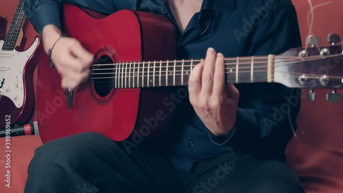 Hands Caucasian man in blue shirt playing red acoustic guitar. The man right hand is wearing a bracelet. Microphone and electric guitar in background photo