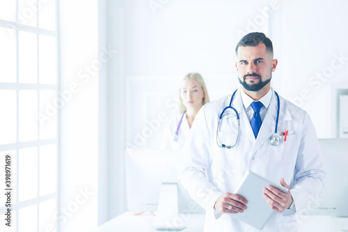 Confident smiling doctor posing and looking at camera with arms crossed, medical staff working on the background