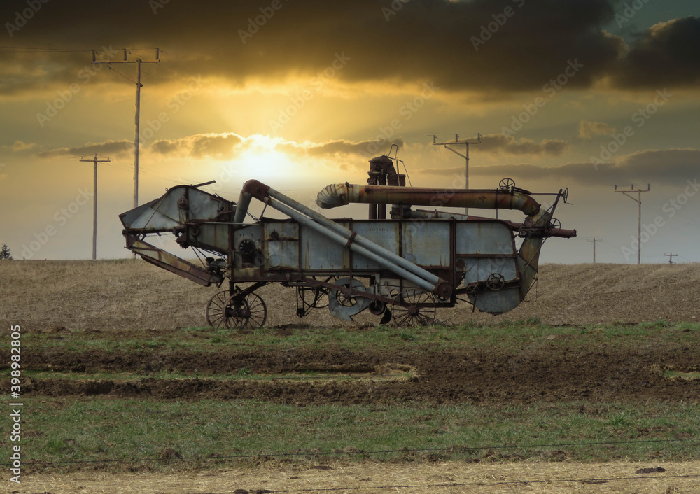 A horizontal photo of a old vintage thrashing machine standing in the field with a beautiful sunset.