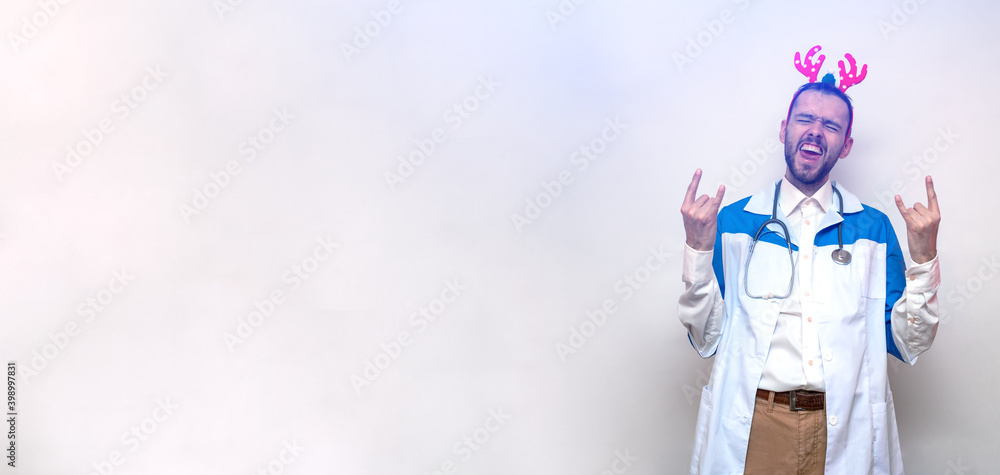 Young doctor with toy deer antlers shows a rocks or yo or yeah sign with his hands. White background. Isolated. Christmas and New Year concept. Portrait. Copy space. Place for text Young doctor with toy deer antlers shows a rocks or yo or yeah sign with his hands. White background. Isolated. Christmas and New Year concept. Portrait. Copy space. Place for text