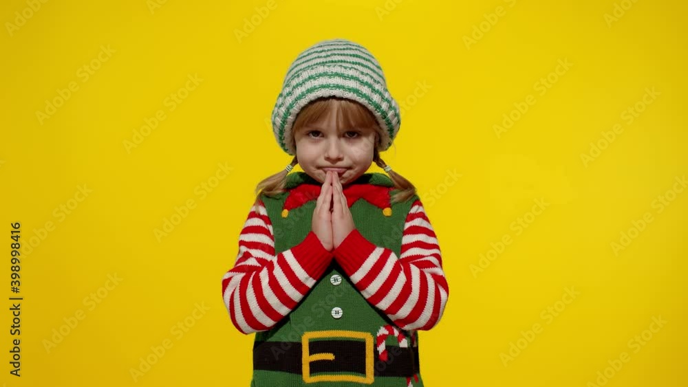 Pleading kid in Christmas elf Santa helper costume begs, prays for forgiveness feels sorry and guilty, keeps palms together, standing over yellow background. Child girl pleads about gifts on New Year Pleading kid in Christmas elf Santa helper costume begs, prays for forgiveness feels sorry and guilty, keeps palms together, standing over yellow background. Child girl pleads about gifts on New Year