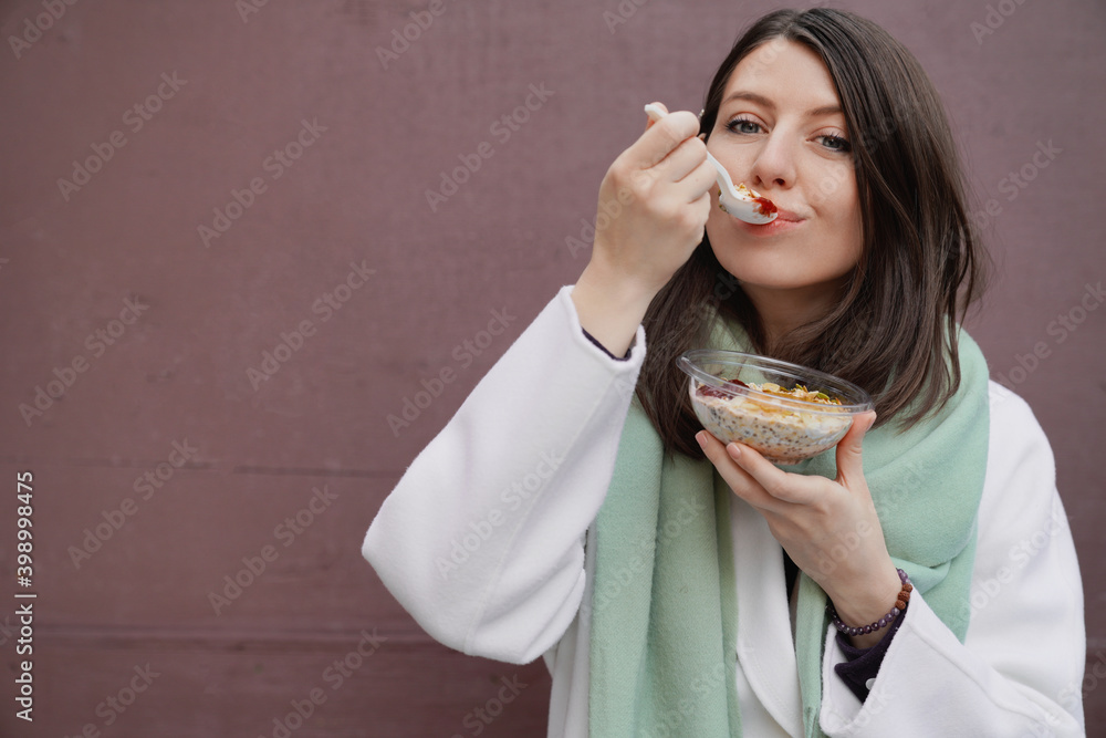 Attractive brown hair young girl in white coat and scarf having breakfast, lunch to-go, cheerful, smiling. Plain wall on background, copy space for text Attractive brown hair young girl in white coat and scarf having breakfast, lunch to-go, cheerful, smiling. Plain wall on background, copy space for text