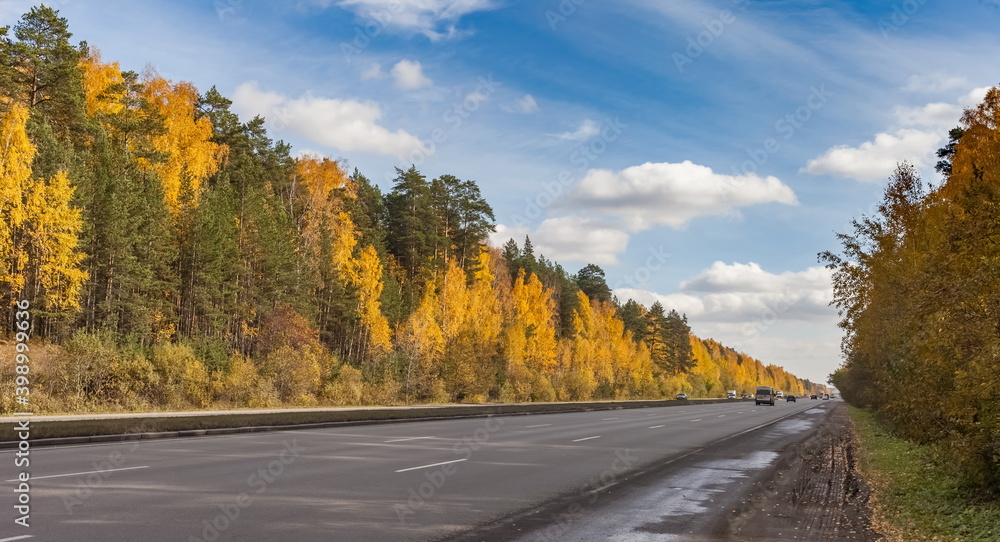 Road with cars on the background of yellow and green trees and blue sky with white clouds in autumn Road with cars on the background of yellow and green trees and blue sky with white clouds in autumn
