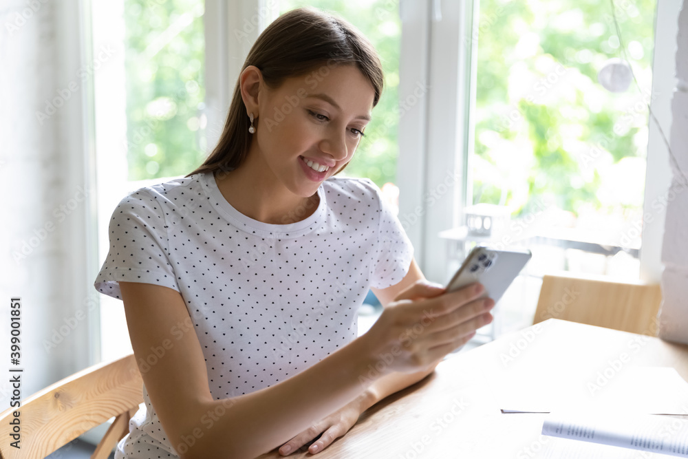 Fototapeta premium Close up smiling young woman holding phone, looking at screen, chatting online with friends, browsing mobile device apps, shopping, reading message in social network, sitting at desk at home