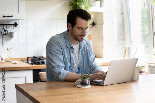 Confident young man wearing glasses using laptop, working, sitting at wooden table in kitchen, checking email in morning, chatting with colleagues in social network, searching information