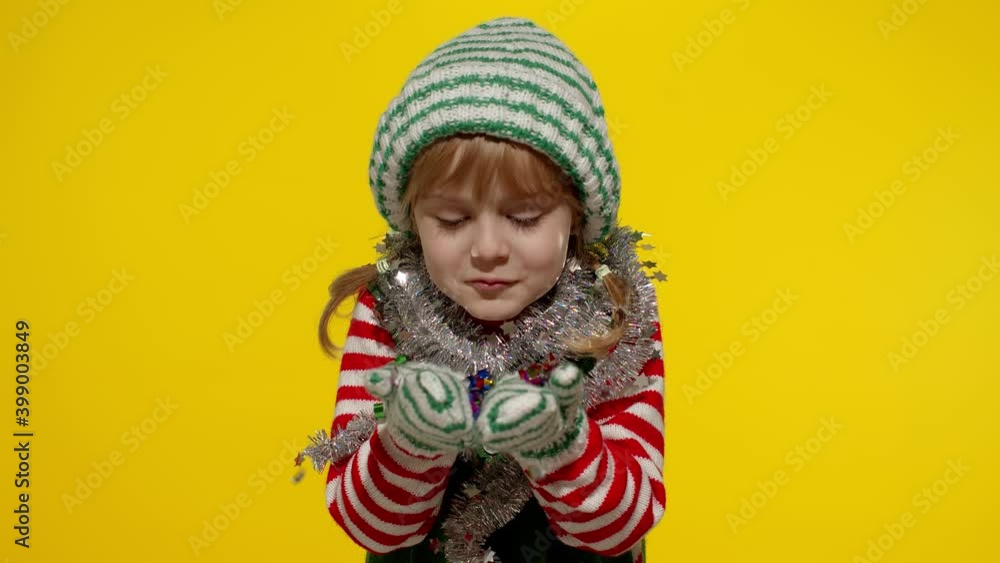 Kid girl in Christmas elf Santa helper costume having fun blows confetti to the camera isolated on yellow background. Child blowing confetti, fooling around. People New Year holidays celebration Kid girl in Christmas elf Santa helper costume having fun blows confetti to the camera isolated on yellow background. Child blowing confetti, fooling around. People New Year holidays celebration
