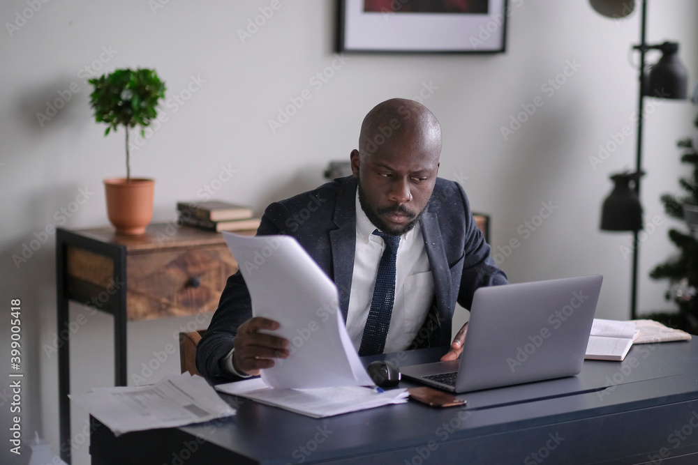 Young African executive reading documents and working on a laptop while sitting at his desk in an office. soft focus. Young African executive reading documents and working on a laptop while sitting at his desk in an office. soft focus.