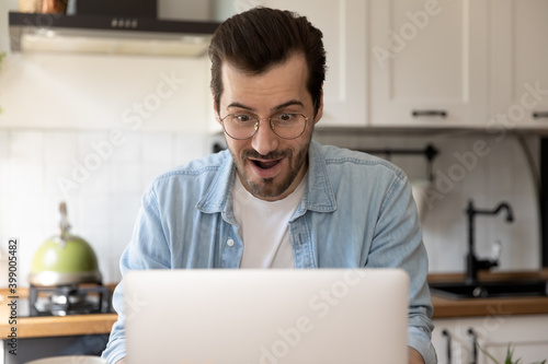 Close up amazed young man wearing glasses using laptop in kitchen, surprised businessman or student with open mouth looking at laptop screen, reading good unexpected news, promotion or lottery win