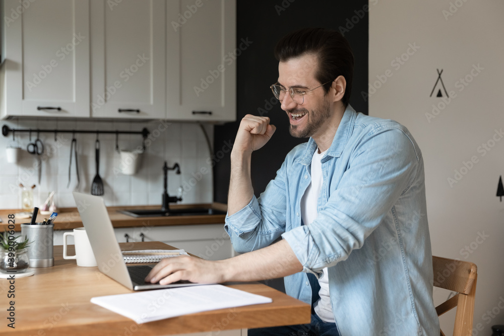 Overjoyed man wearing glasses using laptop in kitchen, showing yes gesture, happy businessman or student looking at computer screen, reading good news, celebrating achievement, money refund Overjoyed man wearing glasses using laptop in kitchen, showing yes gesture, happy businessman or student looking at computer screen, reading good news, celebrating achievement, money refund