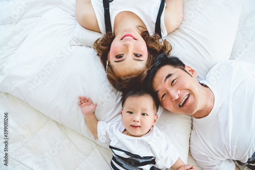 Top view of Asian family with father mother and son on white pillow bed in bedroom at home. Happy parents and child baby concept. People lifestyle in state quarantine from Covid-19 or Coronavirus