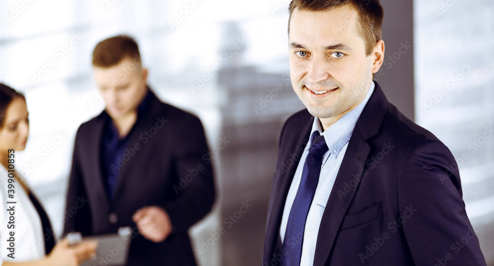 Portrait of a self-confident middle aged businessman in a blue suit, standing in a modern office with his colleagues at the background. Concept of business success Portrait of a self-confident middle aged businessman in a blue suit, standing in a modern office with his colleagues at the background. Concept of business success