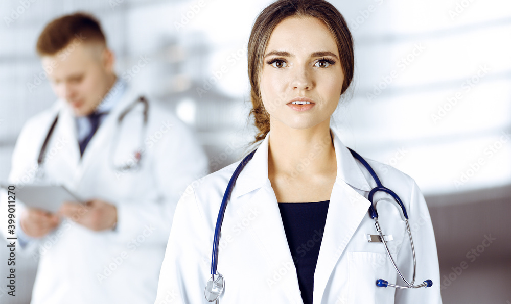 Portrait of a professional friendly woman doctor with a stethoscope, standing with a colleague in a clinic. Young doctors at work in a hospital. Medicine and healthcare concept Portrait of a professional friendly woman doctor with a stethoscope, standing with a colleague in a clinic. Young doctors at work in a hospital. Medicine and healthcare concept