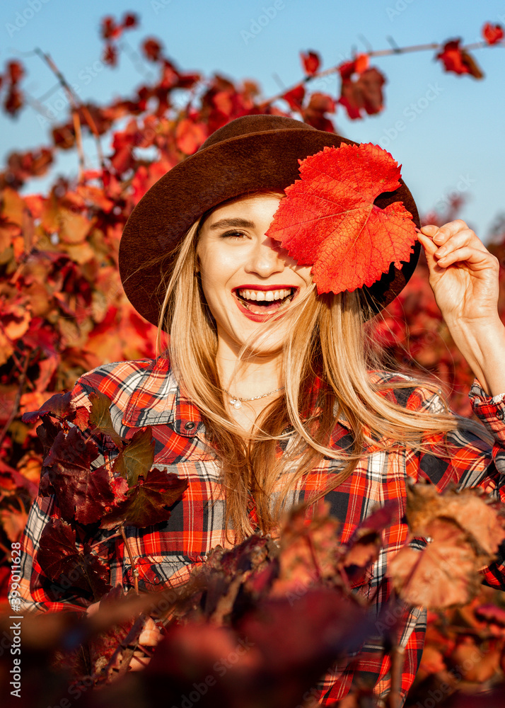 Beautiful young girl in a hat. Behind it is a background of red grape leaves. Autumn photo, a lot of emotions on her face, a grape leaf in her hand, she closes one eye with it and smiles beautifully. Beautiful young girl in a hat. Behind it is a background of red grape leaves. Autumn photo, a lot of emotions on her face, a grape leaf in her hand, she closes one eye with it and smiles beautifully.