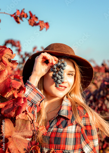 Beautiful young girl in a hat posing for a photographer. Behind it is a background of red grape leaves. Autumn photo  a lot of emotions on your face  a bunch of grapes in your hand