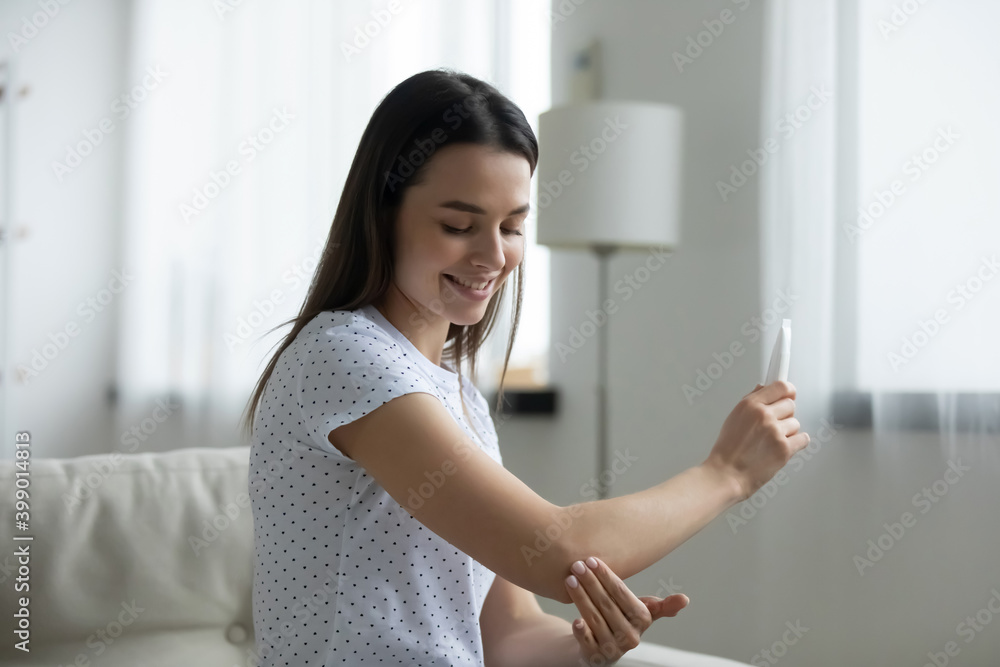 Smiling beautiful young woman applying moisturizing cream on elbow, sitting on cozy couch in living room, happy satisfied attractive girl enjoying smooth healthy skin, daily skincare procedure Smiling beautiful young woman applying moisturizing cream on elbow, sitting on cozy couch in living room, happy satisfied attractive girl enjoying smooth healthy skin, daily skincare procedure