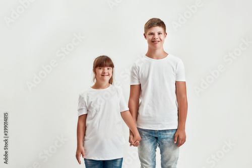 Two kids, disabled boy and girl with Down syndrome smiling at camera, holding each other hands while posing together isolated over white background
