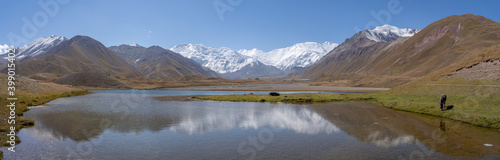 Panoramic view of Achik Tash basecamp of Lenin Peak nowadays Ibn Sina peak in snow-capped Trans-Alay or Trans-Alai mountain range in southern Kyrgyzstan with lake and reflection in foreground